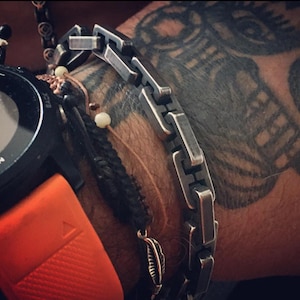 Könnte beinhalten: Nahaufnahme eines Handgelenks mit einem silbernen Kettenarmband, einem schwarzen Perlenarmband mit Federanhänger und einer Garmin-Uhr mit orangefarbenem Armband. Die Uhr zeigt die Uhrzeit und den Batteriestand an.