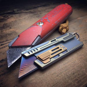 May include: Two utility knives on a wooden surface. One is a red box cutter with a worn appearance. The other is a modern, multi-colored folding knife with a patterned blade and a gold-colored accent.