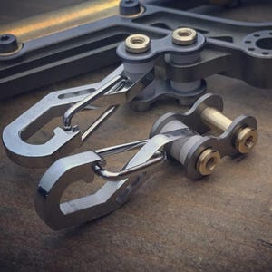 May include: Close-up of several metal carabiner clips and mechanical components. The clips are silver with a curved design, and the other components have gold and gray accents. The items are arranged on a wooden surface.
