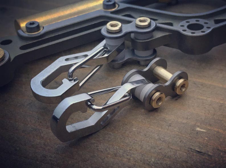 May include: Close-up of several metal carabiner clips and mechanical components. The clips are silver with a curved design, and the other components have gold and gray accents. The items are arranged on a wooden surface.