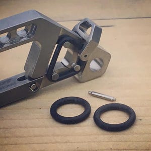 May include: A close-up of a silver metal tool with a hinged design, featuring a series of holes and a circular opening. Two black rubber O-rings and a small silver pin are also present. The items are on a light brown surface.