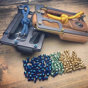 May include: Two metal wallets with leather and rope details, one black and one brown. A collection of blue, green, and gold screws are arranged below. The wallets have a minimalist design with visible screws and metal frames.