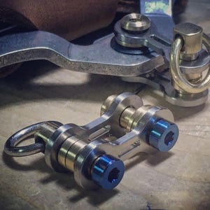 Puede incluir: Primer plano de un llavero metálico con un diseño complejo. El llavero presenta componentes plateados, dorados y azules, incluyendo un anillo y varias piezas de conexión. El diseño parece industrial o mecánico.