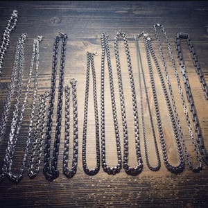 May include: An assortment of silver-coloured necklaces featuring diverse chain designs, such as ball, box, and curb chains. The necklaces are presented on a wooden surface, displaying various lengths and styles of jewellery.