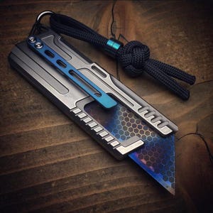 Peut inclure: Un couteau utilitaire argenté et bleu avec un motif hexagonal sur la lame. Le couteau a un accent bleu et une longe noire attachée à un anneau métallique. La lame est partiellement déployée.