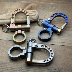 May include: Three metal carabiner clips in gold, blue, and silver tones. The carabiners have a D-shaped design with a hinged gate and a small ring for attaching keys or other items. The background is a wooden surface.
