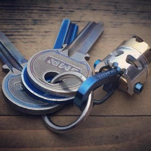 Può includere: Un insieme di chiavi e un portachiavi con una combinazione di colori blu e argento. Le chiavi presentano dettagli blu e il portachiavi riporta la scritta "TITAN". Gli oggetti sono disposti su una superficie di legno.