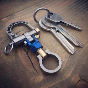 May include: A silver metal key ring with a blue and gold accent, next to a set of keys on a wooden surface. The key ring has a unique design with a carabiner-style clasp. The keys are attached to a silver ring.