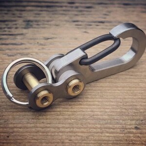 May include: A silver-colored keychain with a black clasp and gold-colored accents. The keychain is made of metal and has a ring for keys. The design is industrial and modern, suitable for everyday use.