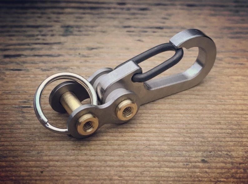 May include: A silver-colored keychain with a black clasp and gold-colored accents. The keychain is made of metal and has a ring for keys. The design is industrial and modern, suitable for everyday use.