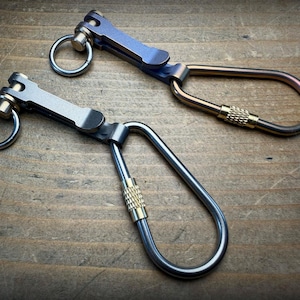 Pode incluir: Dois porta-chaves de metal com conector giratório, argola circular e mosquetão alongado com porca de bloqueio em latão serrilhado. Um tem acabamento cinzento mate, enquanto o outro apresenta um gradiente azul e bronze anodizado a quente.