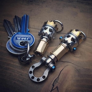 May include: Two custom keychains with a skull design, featuring silver and gold metal accents, and a blue key. The keychains have a unique, industrial aesthetic. The keys are attached to a wooden surface.