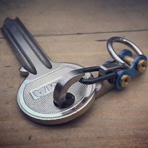 May include: A silver-colored key-shaped keychain with a textured surface. The keychain has a ring and a blue and gold-colored connector. The key has the letters "MA" engraved on it.