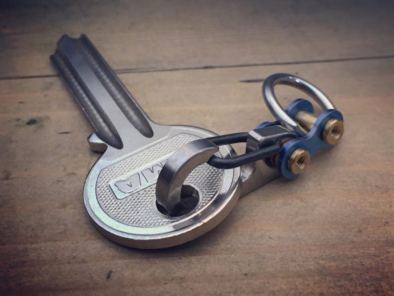 May include: A silver-colored key-shaped keychain with a textured surface. The keychain has a ring and a blue and gold-colored connector. The key has the letters "MA" engraved on it.
