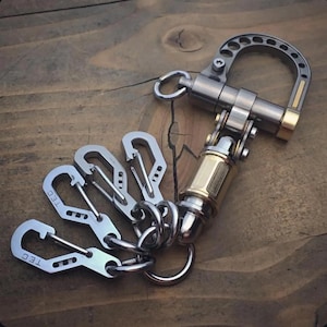 May include: A multi-piece keychain with a silver and gold finish. The keychain features a large, curved clasp, a swivel mechanism, and three smaller carabiner-style clips. The clips have the letters "TEC" engraved on them.