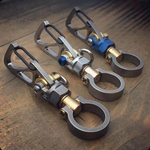 May include: Three metal keychains with a silver finish, brass accents, and a blue bolt. Each keychain features a D-ring and a spring-loaded clasp. The keychains are displayed on a wooden surface.