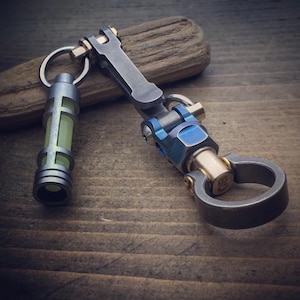 Könnte beinhalten: Ein maßgefertigter Schlüsselanhänger mit einem Metallclip und einem kleinen zylindrischen Licht. Der Schlüsselanhänger weist eine Kombination aus silbernen, blauen und goldfarbenen Metallkomponenten auf. Das Licht strahlt ein sanftes, grünes Leuchten aus. Das Design ist aufwendig und industriell.