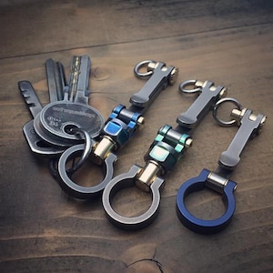 Pode incluir: Porta-chaves com um design moderno. Os porta-chaves apresentam detalhes prateados, dourados e azuis ou verdes. Fabricados em metal, exibem uma construção robusta. Chaves também são visíveis.