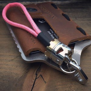 Peut inclure: Un porte-clés multifonction avec une lanière rose, un portefeuille en cuir marron et un outil en métal argenté et doré. Le portefeuille porte le texte "TRAJAN" et "IN USA". Le porte-clés a un porte-clés attaché.