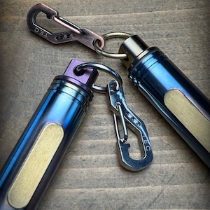 Könnte beinhalten: Zwei zylindrische Metallkapseln mit blau und violett eloxierten Oberflächen, die jeweils eine messingfarbene rechteckige Einlage aufweisen. Beide Kapseln sind an kleinen Metallkarabinern mit der Aufschrift TEC und Spaltringen befestigt.
