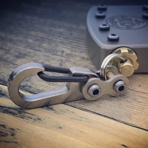 May include: A close-up of a metal carabiner with a black rubber insert, connected to a metal clasp with two screws. The carabiner is silver and the clasp is attached to a gray metal object with screws and a gold-colored knob.