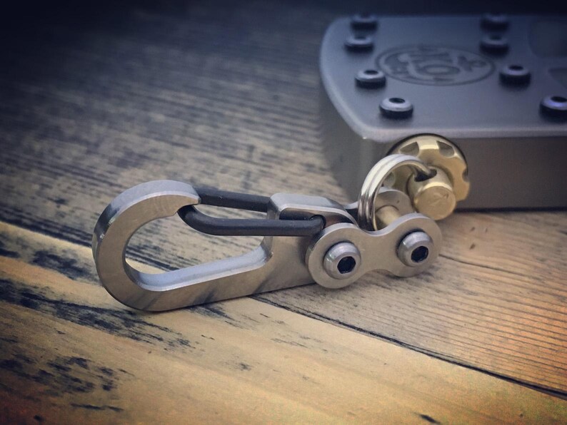 May include: A close-up of a metal carabiner with a black rubber insert, connected to a metal clasp with two screws. The carabiner is silver and the clasp is attached to a gray metal object with screws and a gold-colored knob.