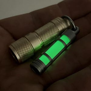 以下が含まれることがあります： 2つの小さな懐中電灯。1つはテクスチャ加工されたメタリックゴールド色で、もう1つは暗いグレーで、緑色に光る中心部があります。どちらもキーリングが付いています。