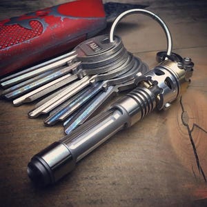 Pode incluir: Um conjunto de chaves em um chaveiro prateado, com um chaveiro de metal prateado e dourado em estilo lanterna. Uma faca vermelha e prateada está no fundo. Os itens estão em uma superfície de madeira.