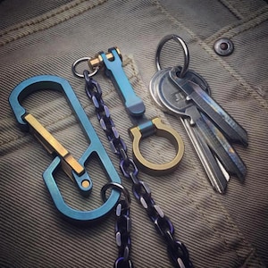May include: A collection of keychains and keys in various metallic finishes. The keychains include a blue carabiner with a gold accent, a blue and gold clasp, a gold ring, and a chain. Several keys are attached to a silver ring.