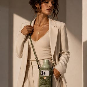 Op de afbeelding: Een olijfgroene gehaakte crossbody flessenhouder met een voorvak voor een smartphone, een bijpassende gevlochten riem en een houten knoopsluiting. De houder bevat een lichtgroene metalen waterfles met een bijpassende dop.