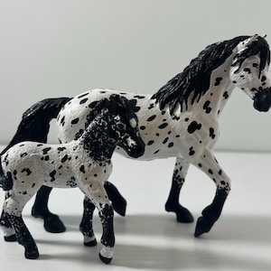May include: A white and black spotted horse figurine with a smaller foal. The adult horse has a black mane and tail. The foal has a similar pattern. The figurines are made of a hard plastic material.