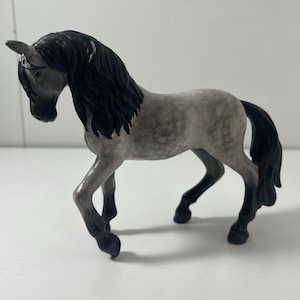 May include: A figurine of a unicorn horse with a gray body and black mane and tail. The unicorn has a single horn and is posed in a walking stance. The figurine is made of a solid material and is painted with detail.