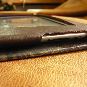 Puede incluir: Una funda de cuero marr&oacute;n para un lector electr&oacute;nico o una tableta. La funda tiene una solapa que cubre la pantalla y un ribete de metal plateado alrededor de los bordes.