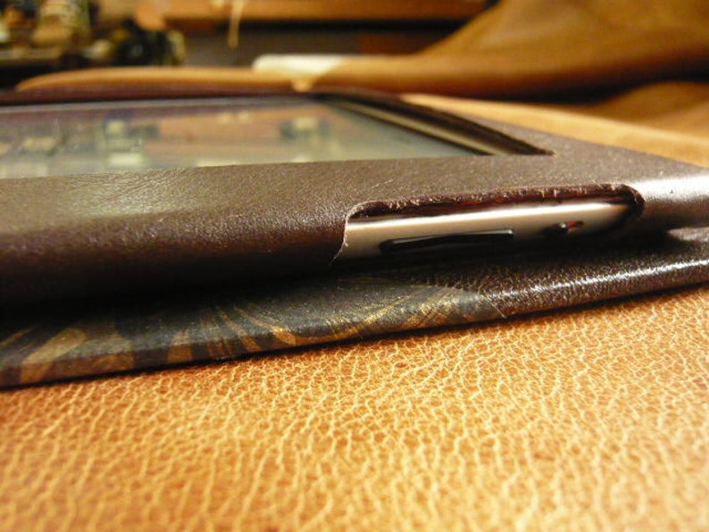 Puede incluir: Una funda de cuero marr&oacute;n para un lector electr&oacute;nico o una tableta. La funda tiene una solapa que cubre la pantalla y un ribete de metal plateado alrededor de los bordes.