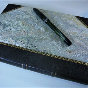 May include: A black leather-bound journal with a marbled paper cover and a gold-colored border. A black pen is lying on top of the journal.
