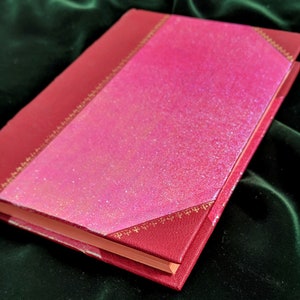 May include: A red hardcover journal with a pink glitter cover and gold accents. The journal is closed and lying on a green surface.