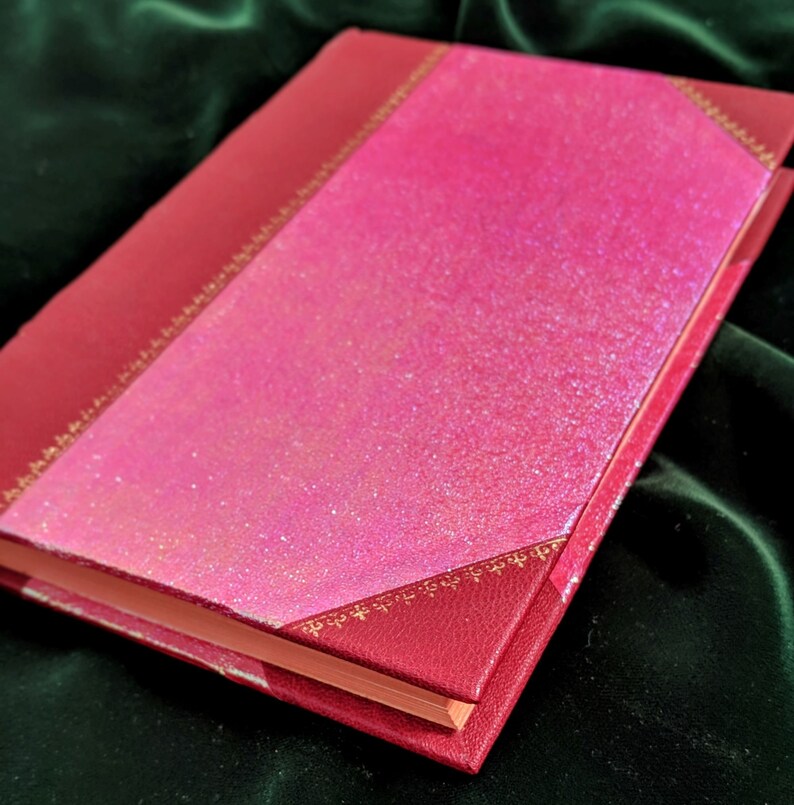 May include: A red hardcover journal with a pink glitter cover and gold accents. The journal is closed and lying on a green surface.