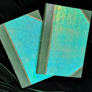 May include: Two green leather-bound journals with a teal iridescent cover. The journals have gold accents and are closed.