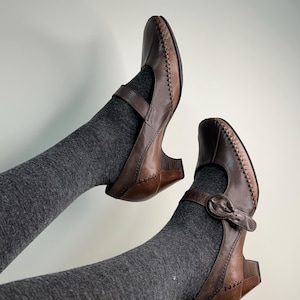 Może przedstawiać: Brązowe sk&oacute;rzane buty Mary Jane z zapięciem na klamrę i małym obcasem. Buty mają ozdobne przeszycia wzdłuż krawędzi i pasek na g&oacute;rze stopy. Buty są noszone z szarymi skarpetami.