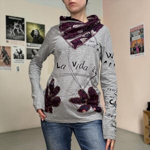 Könnte beinhalten: Graues Langarmshirt mit Rollkragen und floralen Applikationen. Das Shirt trägt den Text "La Vida &" und andere handschriftliche Phrasen. Ein gemusterter Schal in Lila- und Weißtönen wird um den Hals getragen.
