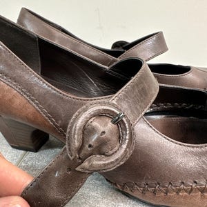 Może przedstawiać: Para brązowych sk&oacute;rzanych but&oacute;w w stylu Mary Jane z zapięciem na klamrę. Buty mają zaokrąglony nosek, mały obcas i ozdobne szwy. Klamra ma okrągły kształt z metalowym zapięciem w kolorze srebrnym. Wysokość obcasa ok. 5 cm.