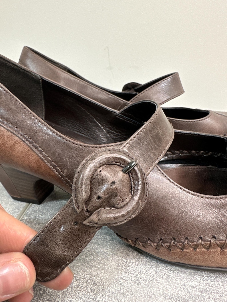 Może przedstawiać: Para brązowych sk&oacute;rzanych but&oacute;w w stylu Mary Jane z zapięciem na klamrę. Buty mają zaokrąglony nosek, mały obcas i ozdobne szwy. Klamra ma okrągły kształt z metalowym zapięciem w kolorze srebrnym. Wysokość obcasa ok. 5 cm.