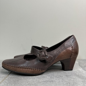 Może przedstawiać: Brązowe sk&oacute;rzane buty w stylu Mary Jane z zapięciem na klamrę i obcasem słupkowym. Buty mają zaokrąglony nosek i ozdobne przeszycia. Wysokość obcasa to około 5 cm.