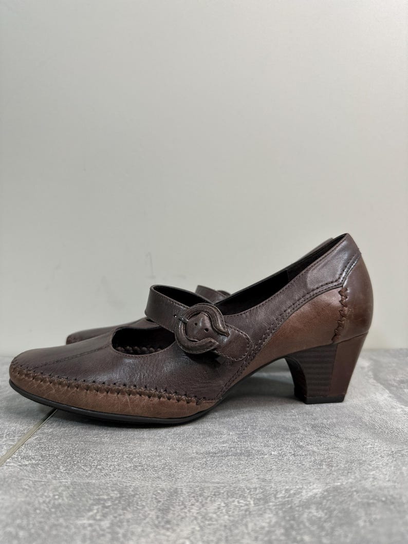 Może przedstawiać: Brązowe sk&oacute;rzane buty w stylu Mary Jane z zapięciem na klamrę i obcasem słupkowym. Buty mają zaokrąglony nosek i ozdobne przeszycia. Wysokość obcasa to około 5 cm.