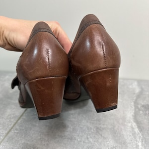 Może przedstawiać: Para brązowych sk&oacute;rzanych but&oacute;w na niskim obcasie. Buty mają zaokrąglony nosek i pasek z klamrą. Obcasy mają około 5 cm wysokości. Buty są używane i noszą ślady użytkowania.