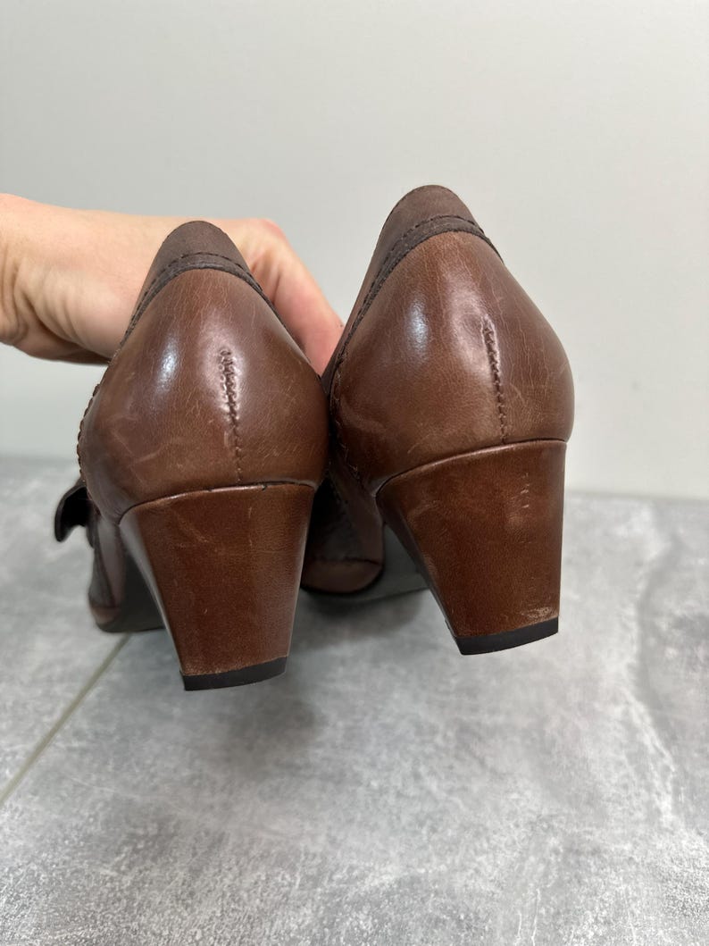 Może przedstawiać: Para brązowych sk&oacute;rzanych but&oacute;w na niskim obcasie. Buty mają zaokrąglony nosek i pasek z klamrą. Obcasy mają około 5 cm wysokości. Buty są używane i noszą ślady użytkowania.