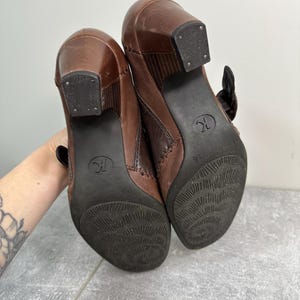 Może przedstawiać: Para brązowych sk&oacute;rzanych botk&oacute;w na obcasie z klamrą. Podeszwy są ciemnobrązowe z teksturowanym wzorem, a obcasy kwadratowe. Buty są w rozmiarze 36.