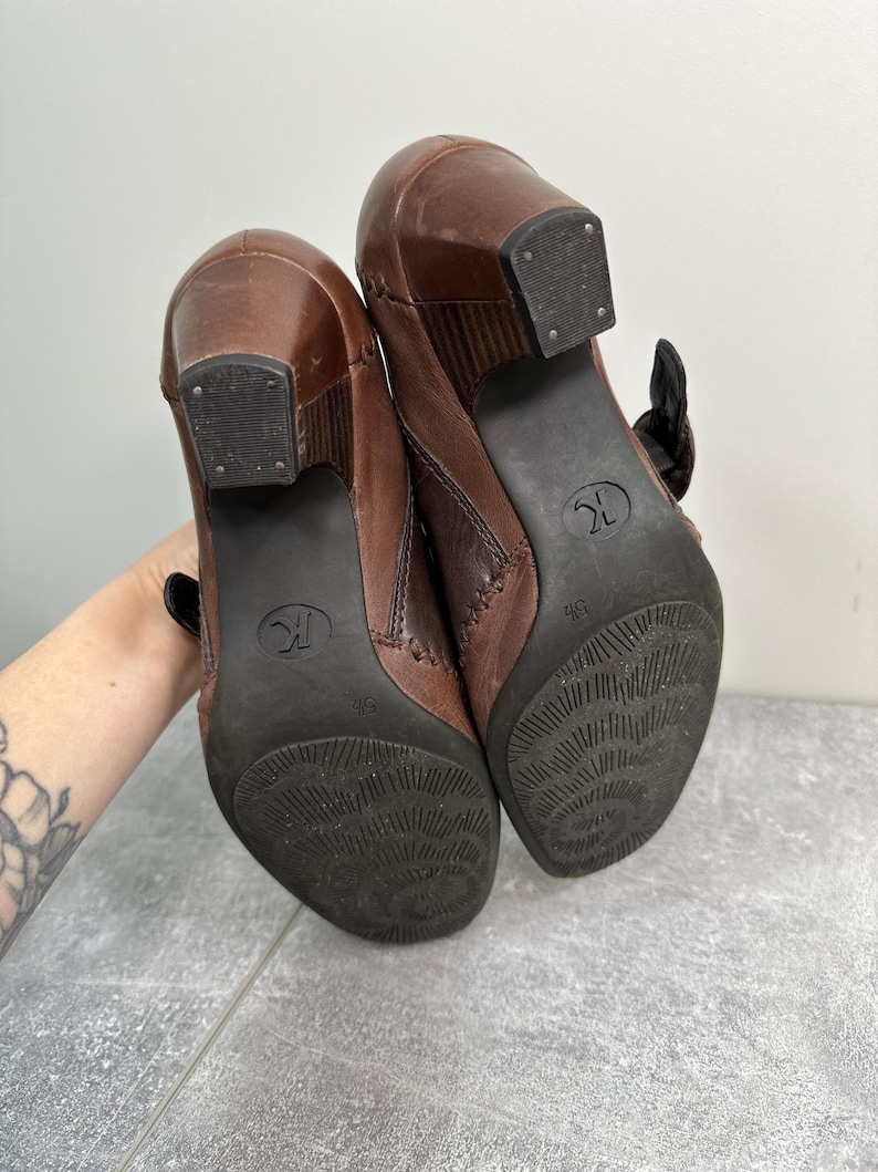 Może przedstawiać: Para brązowych sk&oacute;rzanych botk&oacute;w na obcasie z klamrą. Podeszwy są ciemnobrązowe z teksturowanym wzorem, a obcasy kwadratowe. Buty są w rozmiarze 36.