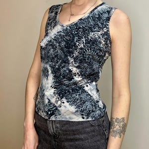 May include: Sleeveless top with a scoop neckline. The top has a black, white, and grey abstract design. The fabric looks to be a soft, lightweight material. The top is worn with black denim jeans.