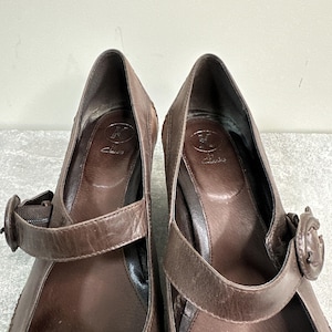 Może przedstawiać: Para brązowych sk&oacute;rzanych but&oacute;w na obcasie w stylu Mary Jane. Buty mają pasek na g&oacute;rze stopy z zapięciem na klamrę. Obcasy są średniej wysokości. Marka "Clarks" jest widoczna na wkładce.